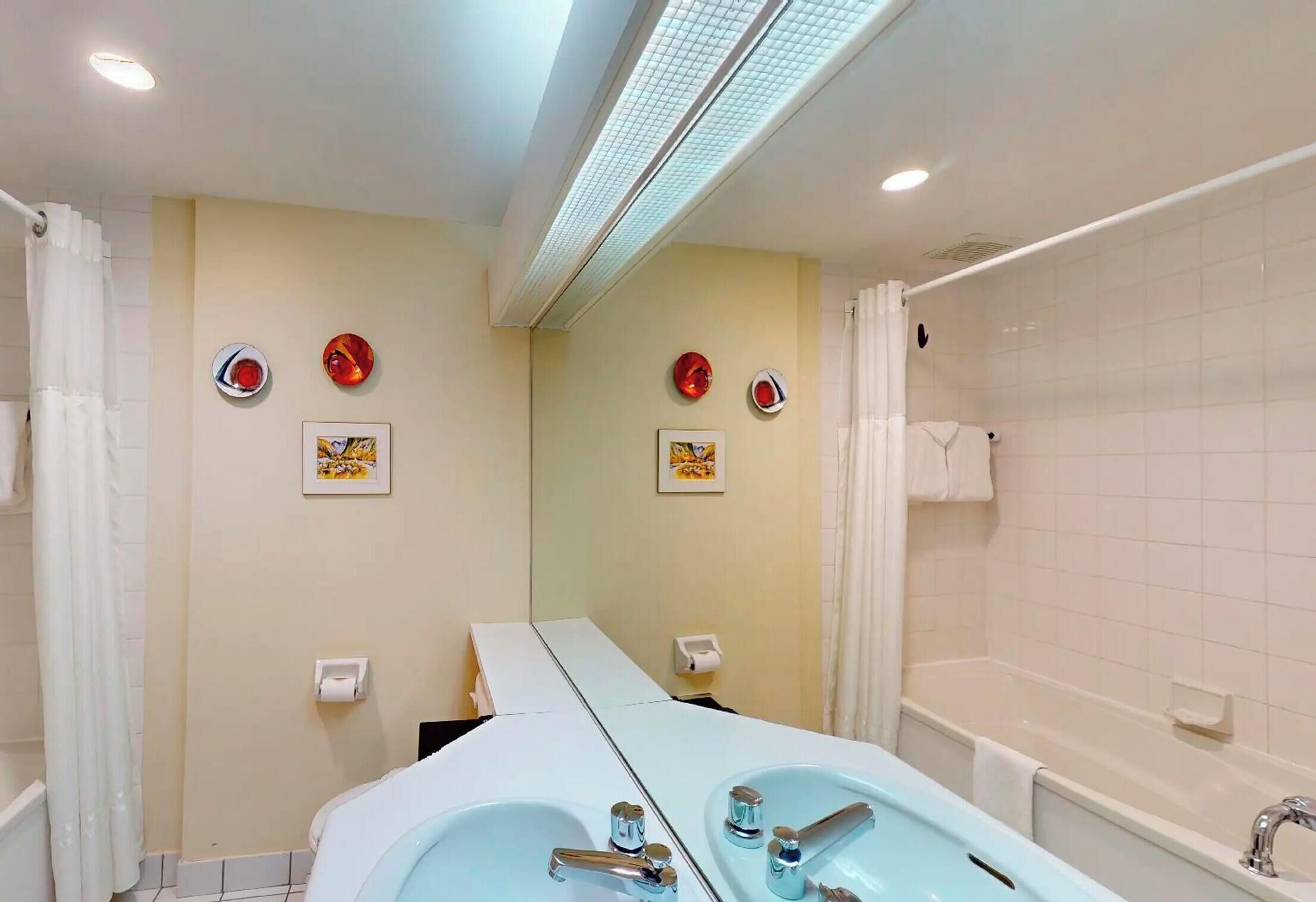 Een badkamer met twee wasbakken, een bad en een douche. Er hangen handdoeken en een toiletrolhouder aan de muur.