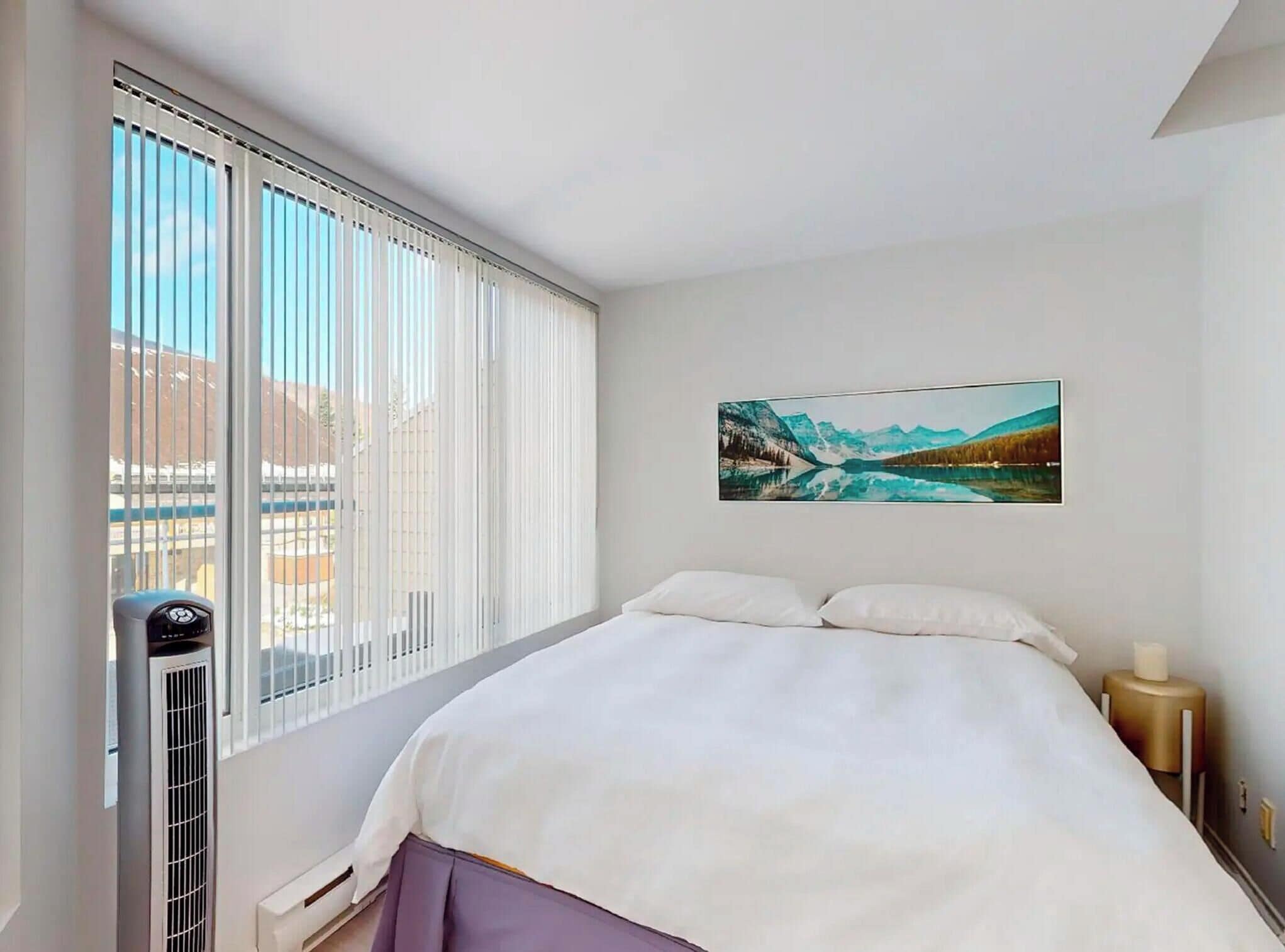 A hotel room with a bed, a window with blinds, a painting on the wall, and a heater.
