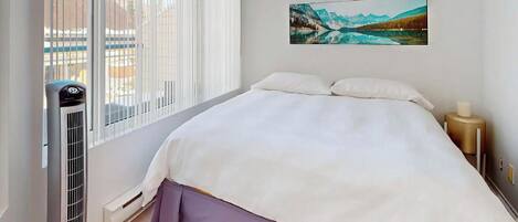 A hotel room with a bed, a window with blinds, a painting on the wall, and a heater.