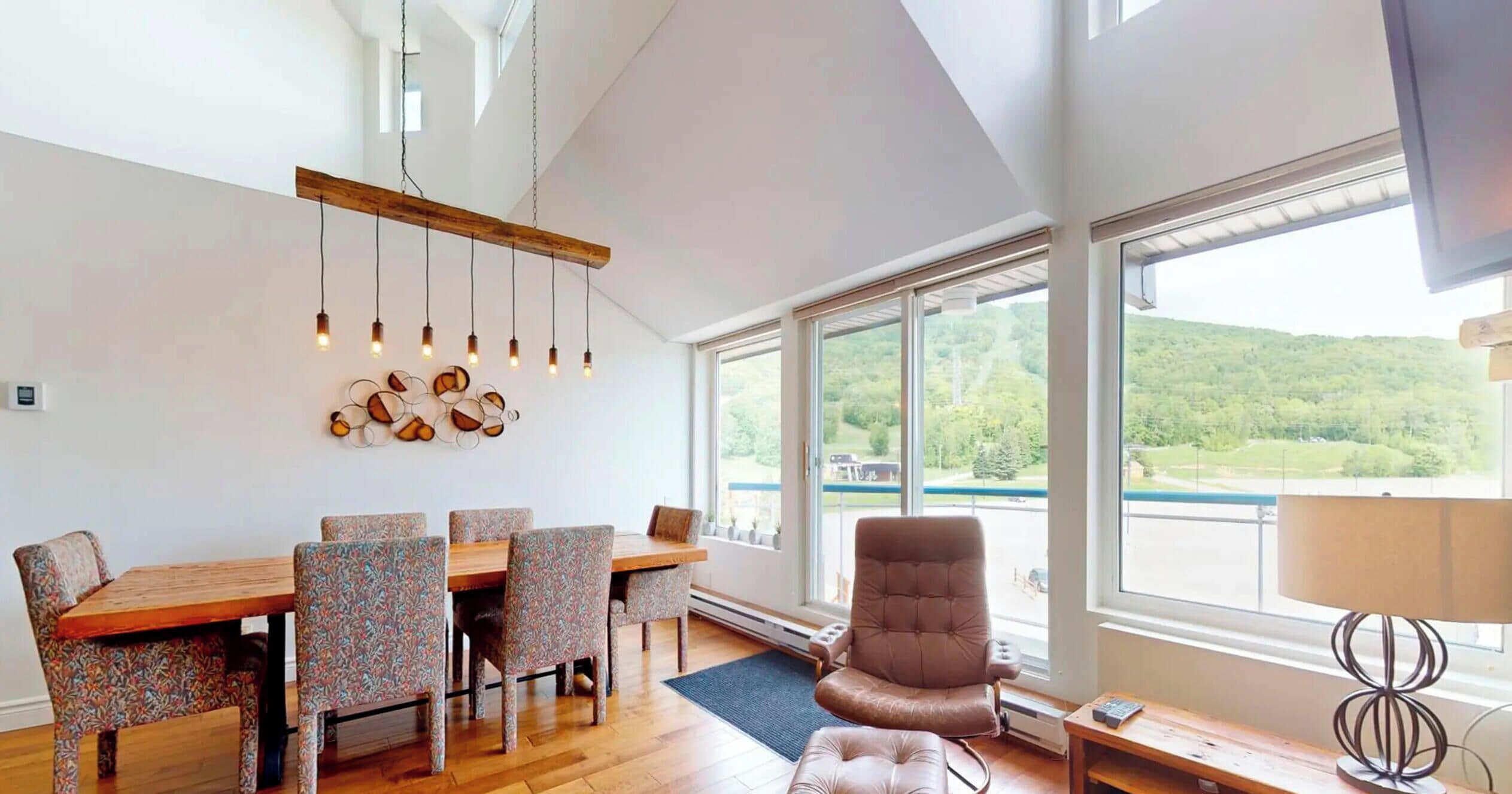 Un comedor moderno con una mesa de madera y sillas estampadas, un ventanal con vistas a la vegetación y una lámpara colgante.