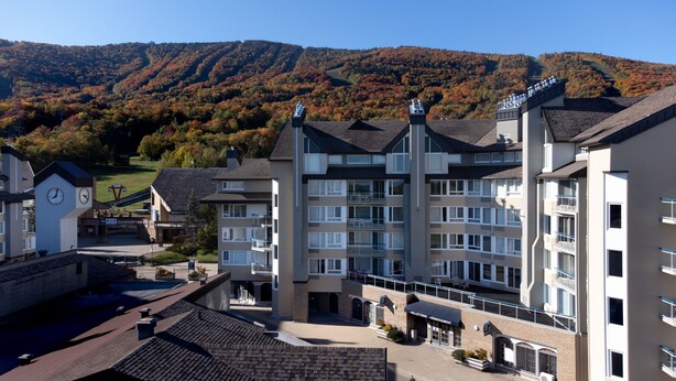 A multi-story hotel with a central courtyard, a clock tower, and a view of autumn-colored trees in the background. - Hébergement Mont-Sainte-Anne (Beaupre)