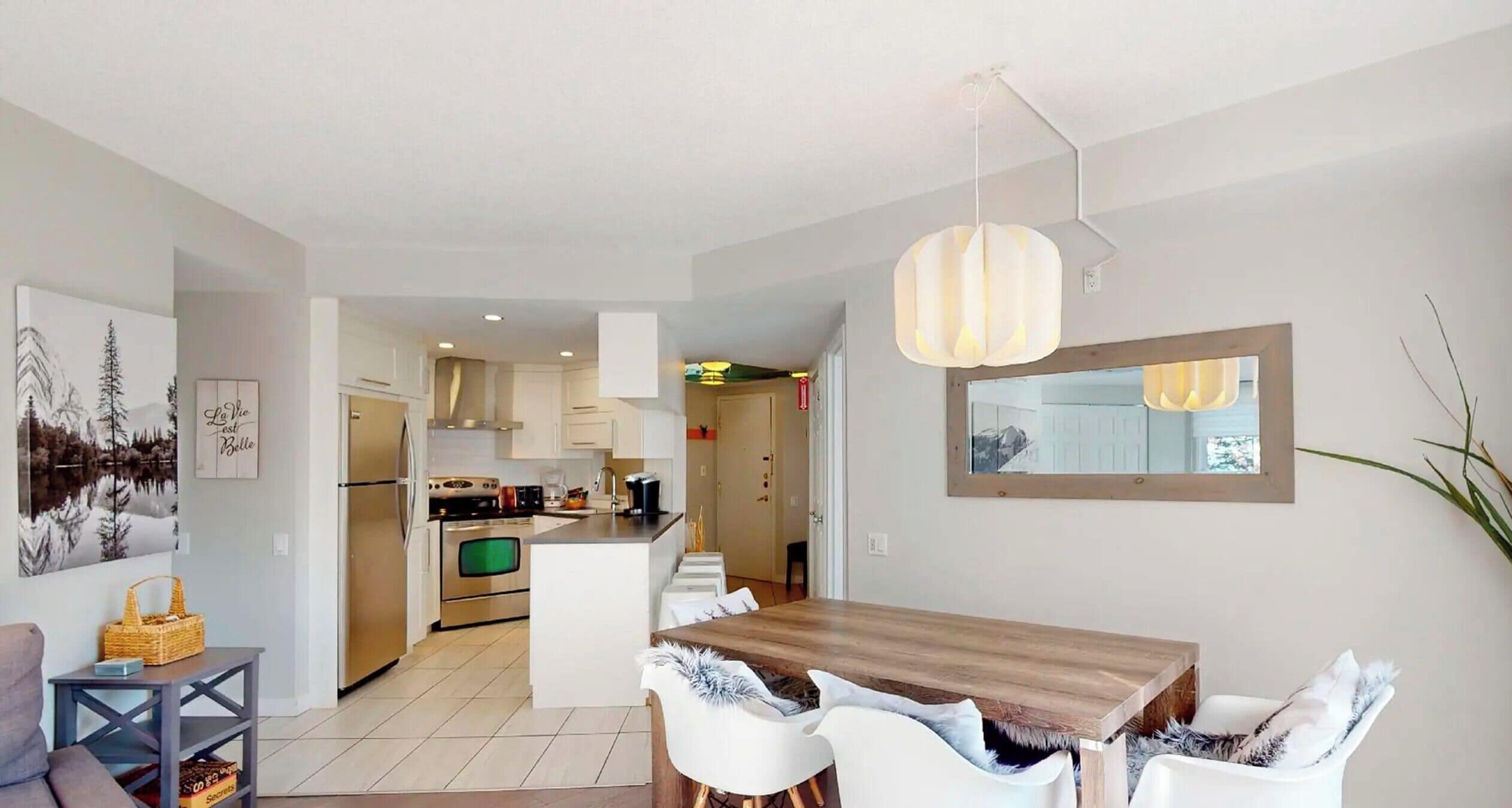 Una cocina moderna con armarios blancos, un comedor con mesa de madera y sillas blancas, y un espejo grande en la pared.