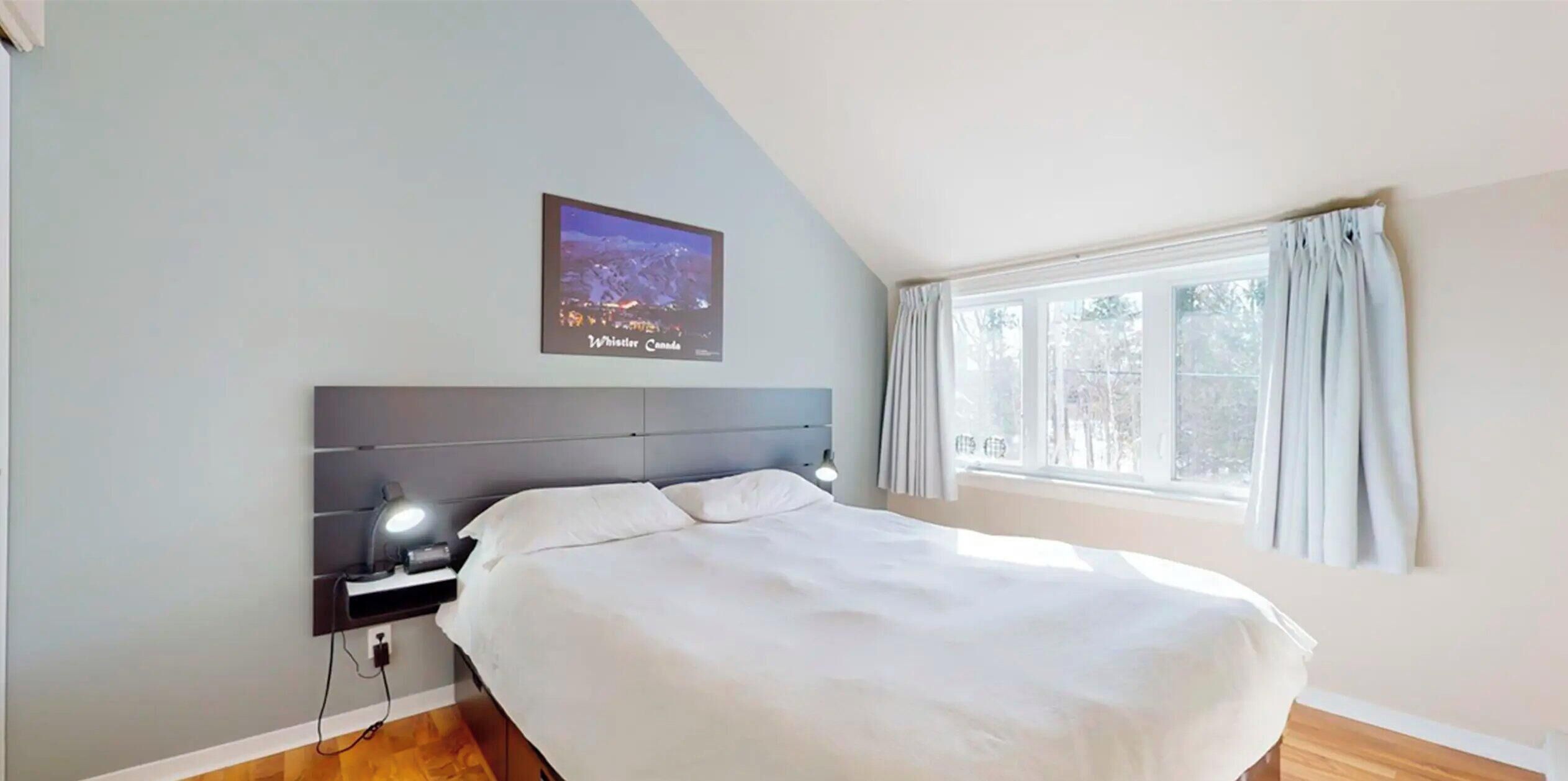 Quarto com cama, mesa de cabeceira, TV na parede e janela com cortinas.