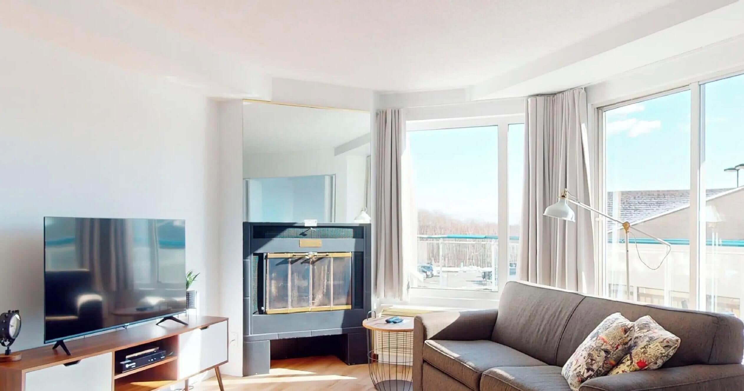 A modern living room with a flat-screen TV, a fireplace, a sofa, and large windows offering a view of the outdoors.