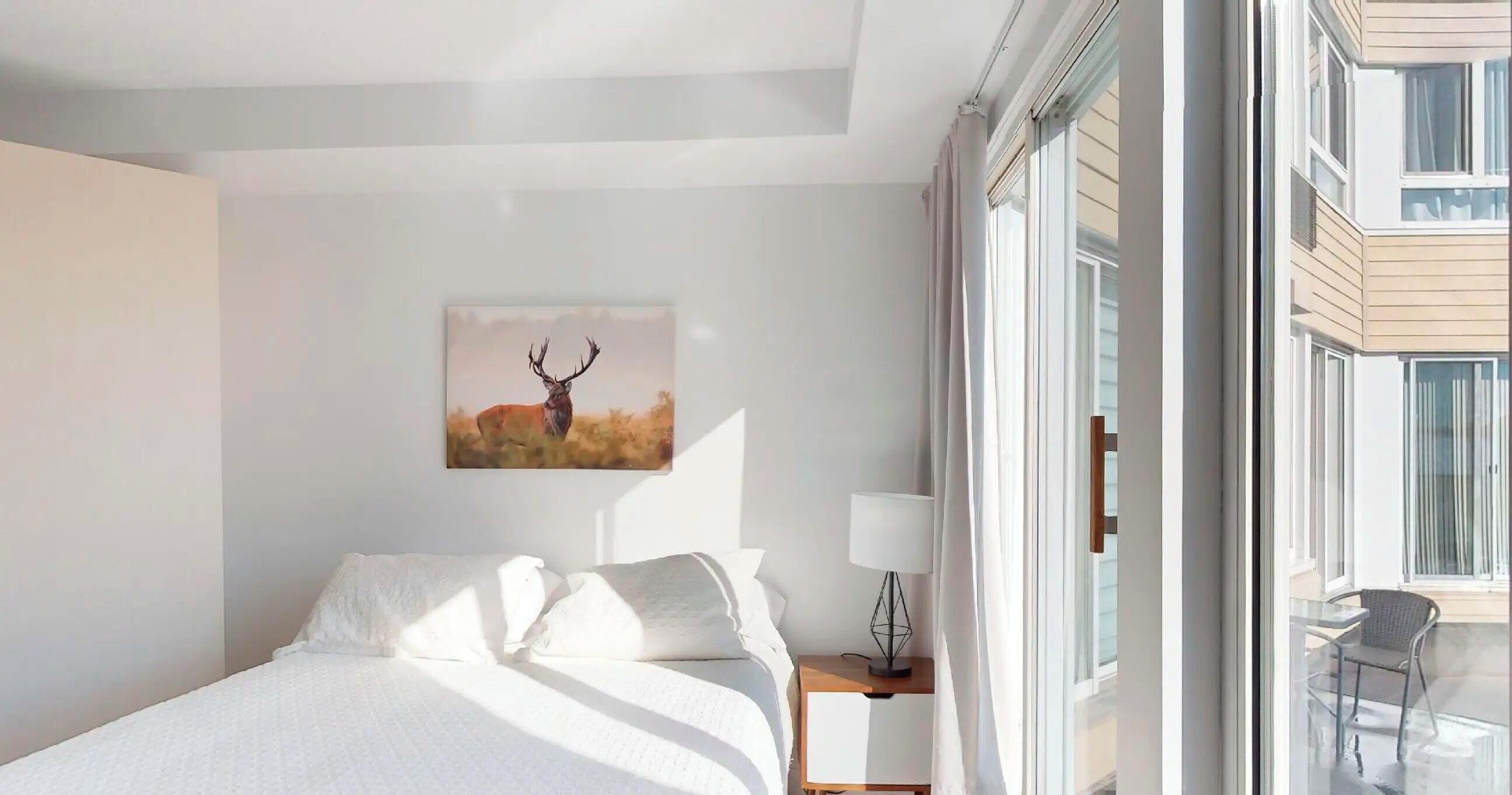 Une chambre d’hôtel avec un lit, une table de chevet avec une lampe, un tableau représentant un cerf et une vue sur le balcon extérieur.
