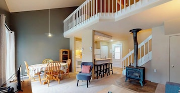 A spacious living area with a high ceiling, a dining table with chairs, a blue armchair, a kitchen area, and a staircase with a white railing.