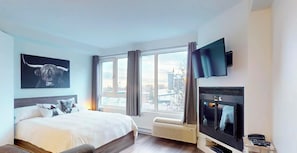 A hotel room with a bed, a mounted TV, a fireplace, and a large window with curtains. - Hébergement Mont-Sainte-Anne (Beaupre)