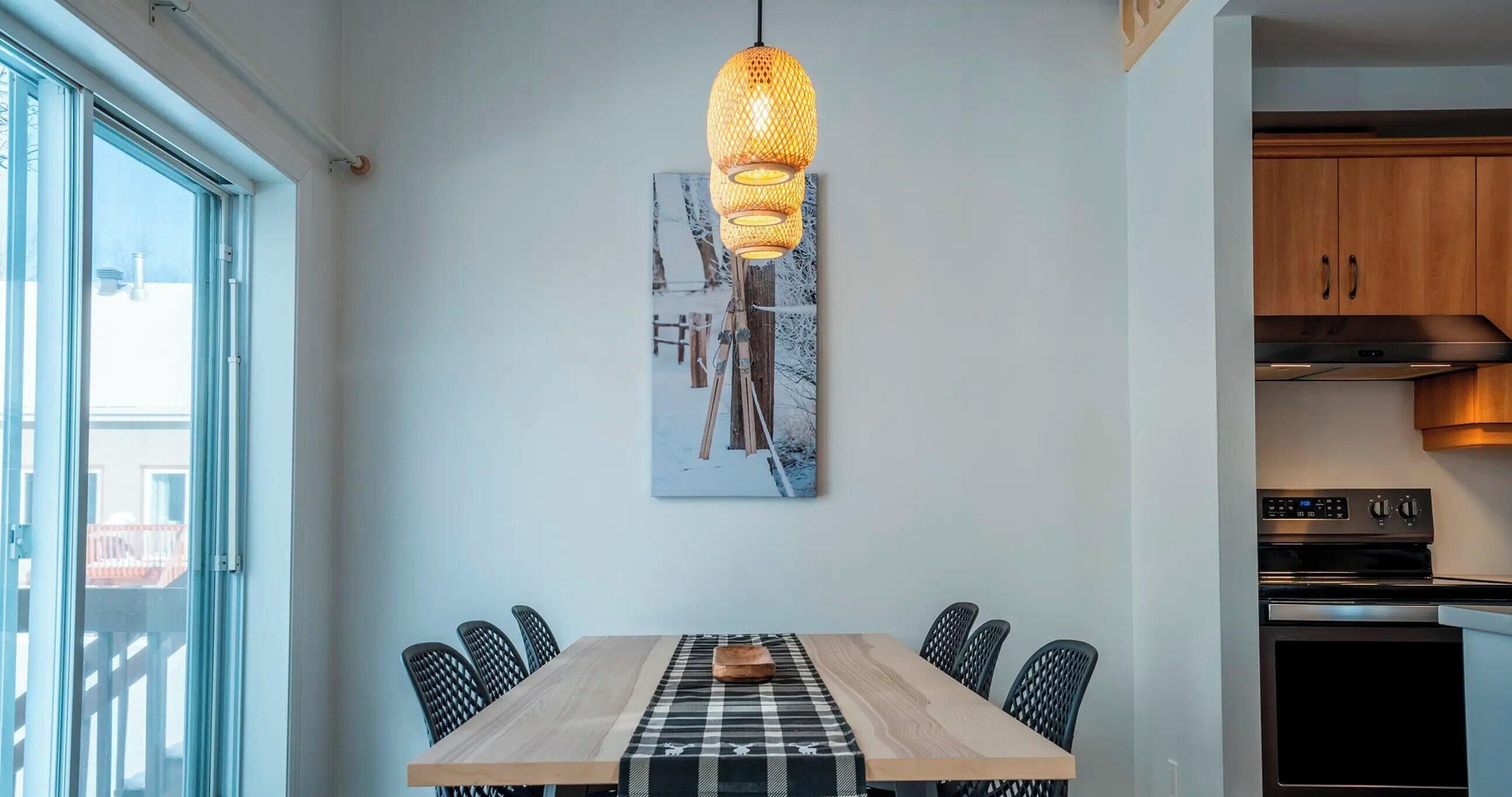 Un coin repas avec une table en bois, une nappe à carreaux noirs et blancs et six chaises. Un luminaire suspendu en fibres tressées est accroché au-dessus.
