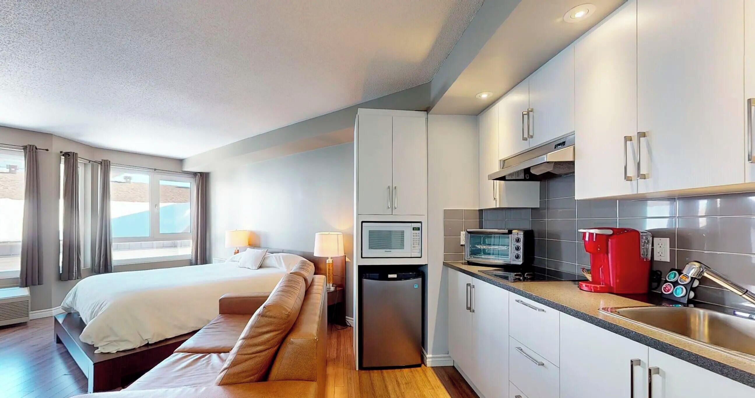 A hotel room with a bed, a sofa, a kitchenette, and a window with curtains.
