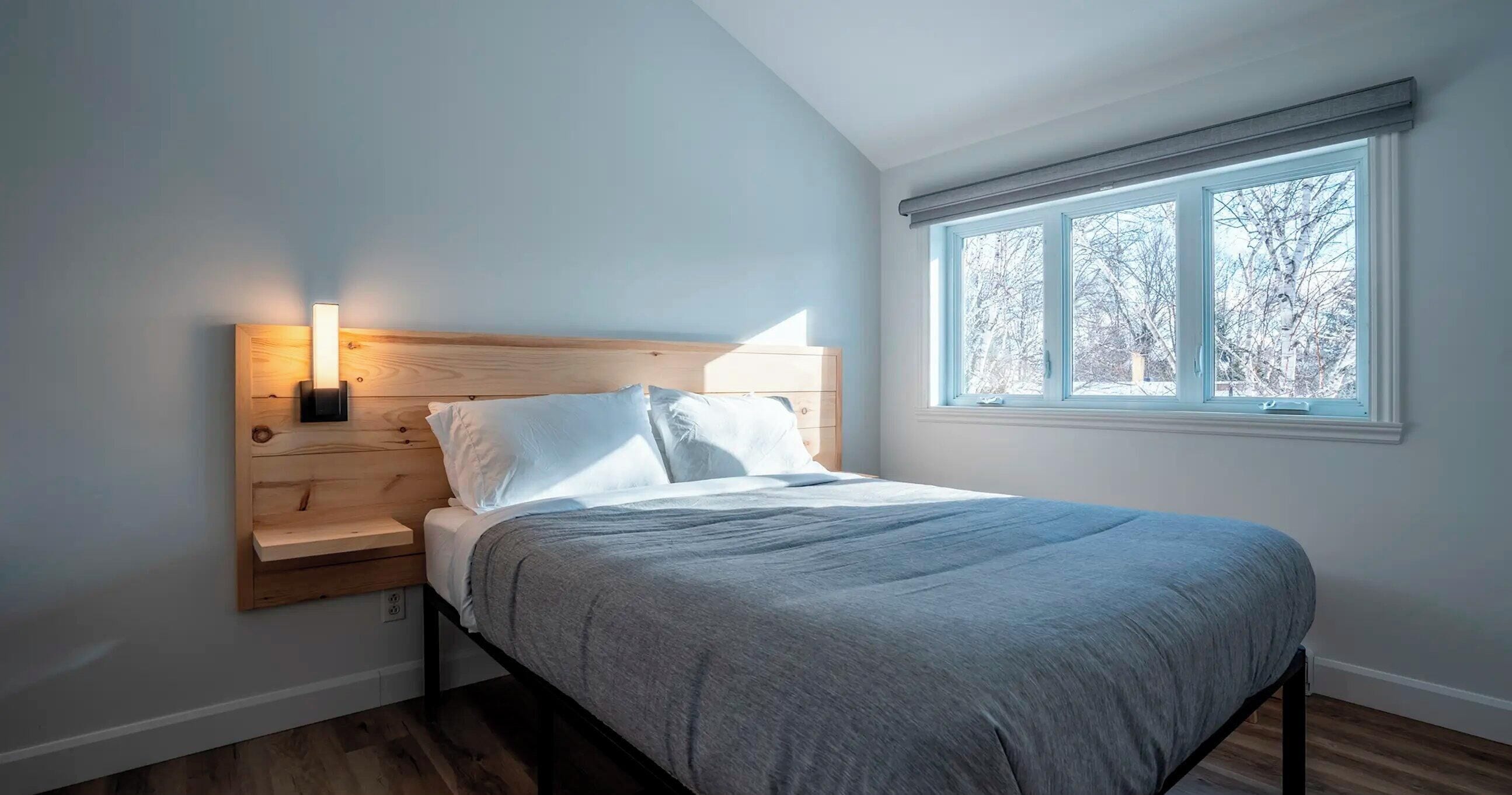 A bedroom with a bed, bedside table, and a window with a view of trees.