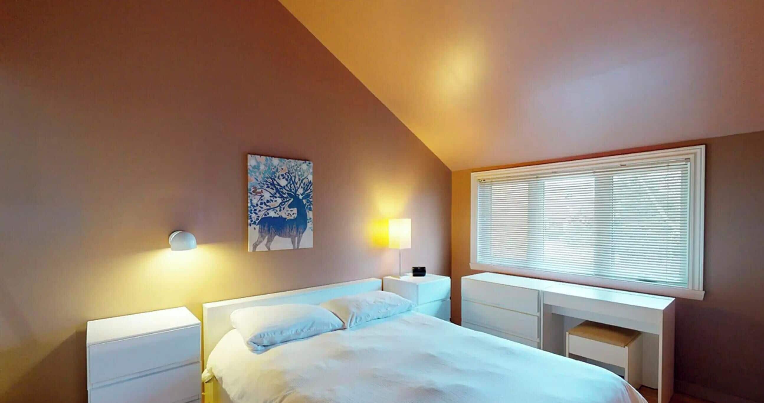 Une chambre à coucher avec un lit, des tables de chevet, une commode, une lampe, un tableau et une fenêtre avec des stores.