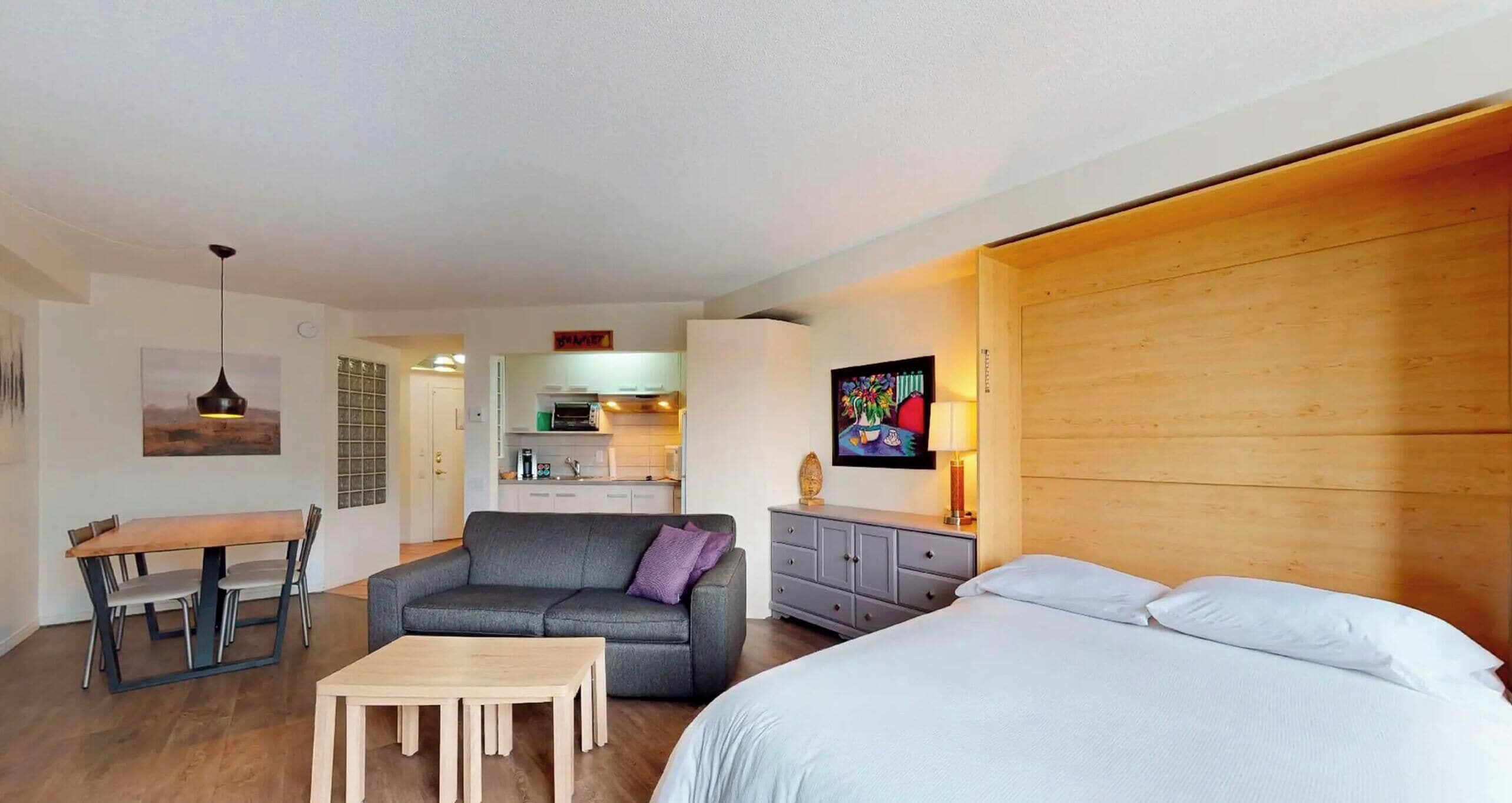 Ein modernes Hotelzimmer mit einem Holzkopfteil, einer Couch, einem Esstisch mit Stühlen, einem kleinen Holztisch und einer Küchenzeile. An der Wand hängt ein Gemälde.