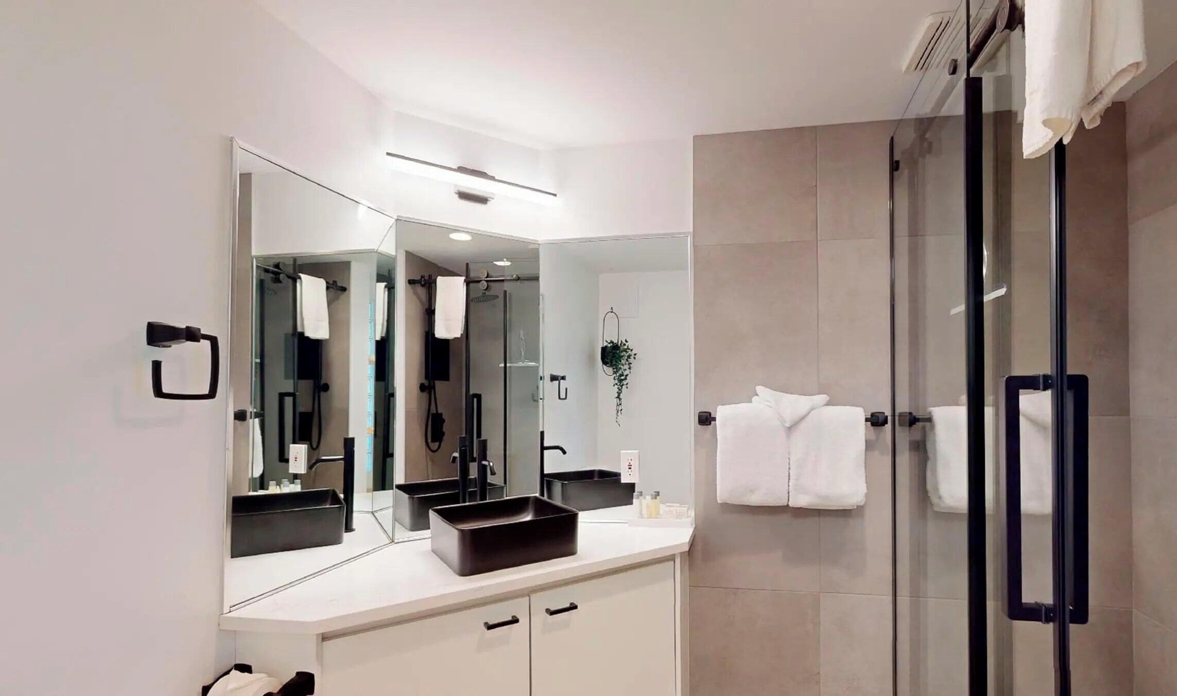 A modern bathroom with a large mirror, a glass shower enclosure, and a white vanity with a black sink.