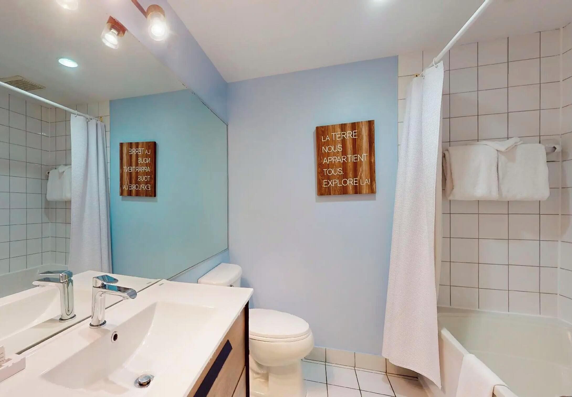 A modern bathroom with a white sink, toilet, and bathtub. There are two wooden signs on the walls with motivational text.