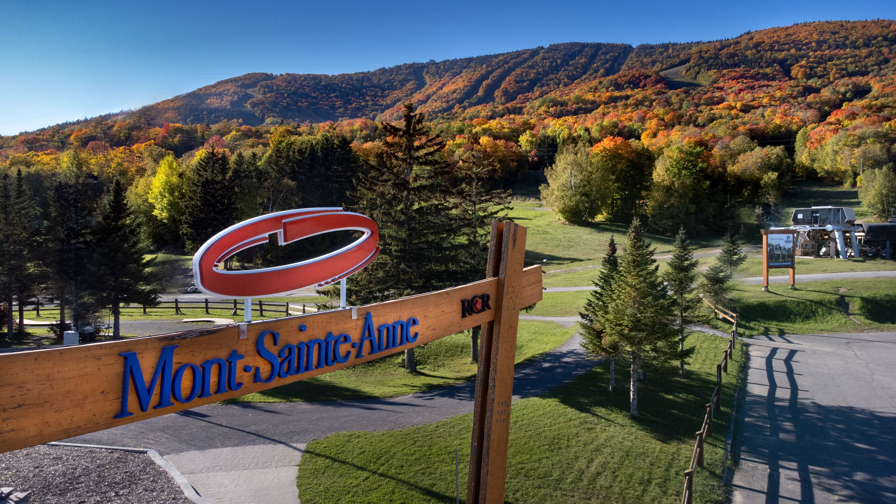 Un panneau en bois sur lequel est inscrit « Mont-Sainte-Anne », entouré d’un paysage pittoresque composé d’arbres aux couleurs automnales et d’une montagne en arrière-plan.