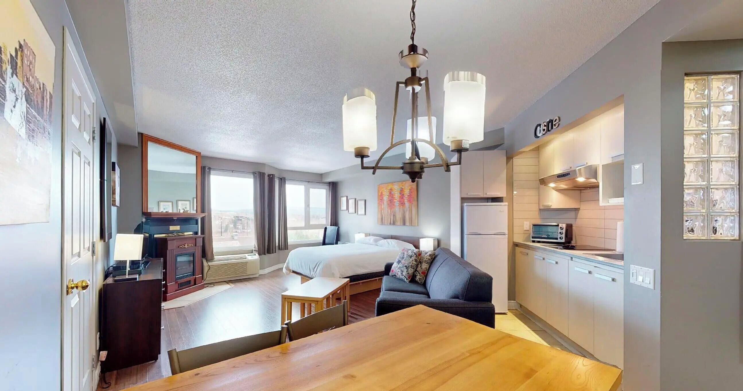 Una habitación de hotel moderna con una mesa de comedor de madera, un sofá, una cama, una kitchenette y un ventanal grande.