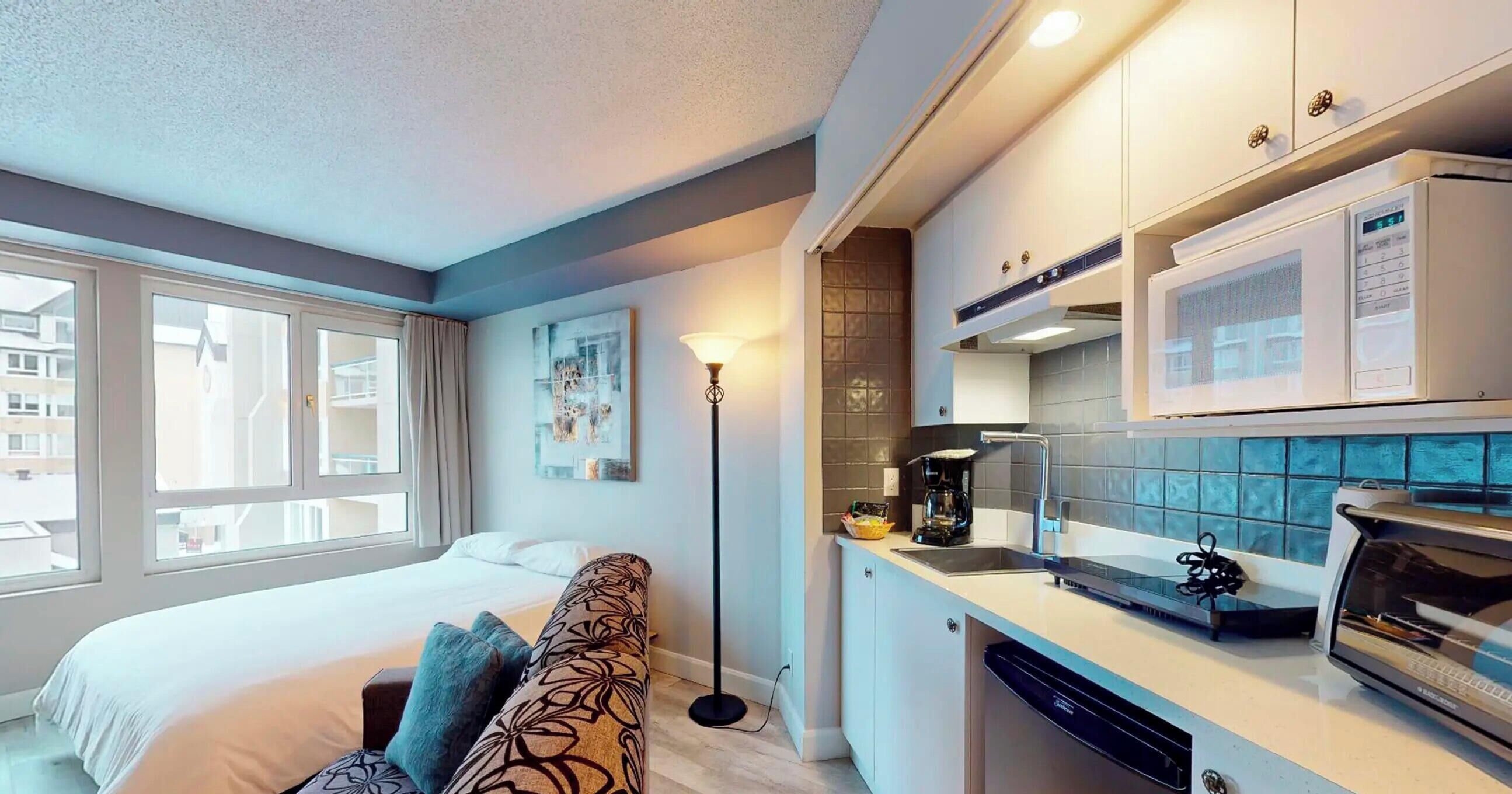 Una habitación de hotel moderna con cama, sofá, lámpara de pie, kitchenette y ventana con vistas a edificios.