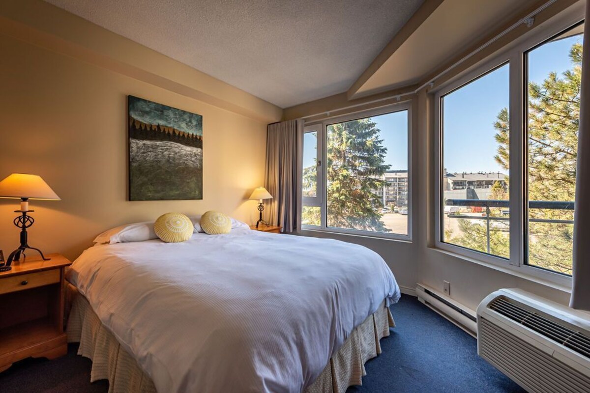A hotel room with a large bed, two bedside lamps, a painting on the wall, and a view of trees and buildings through large windows.