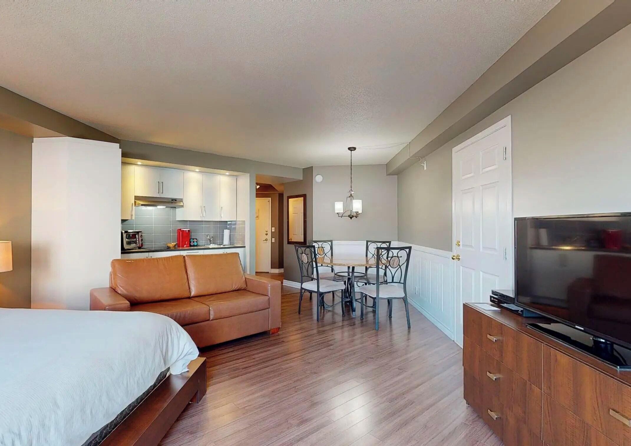 A hotel room with a bed, a sofa, a dining table with chairs, a TV, and a kitchen area.