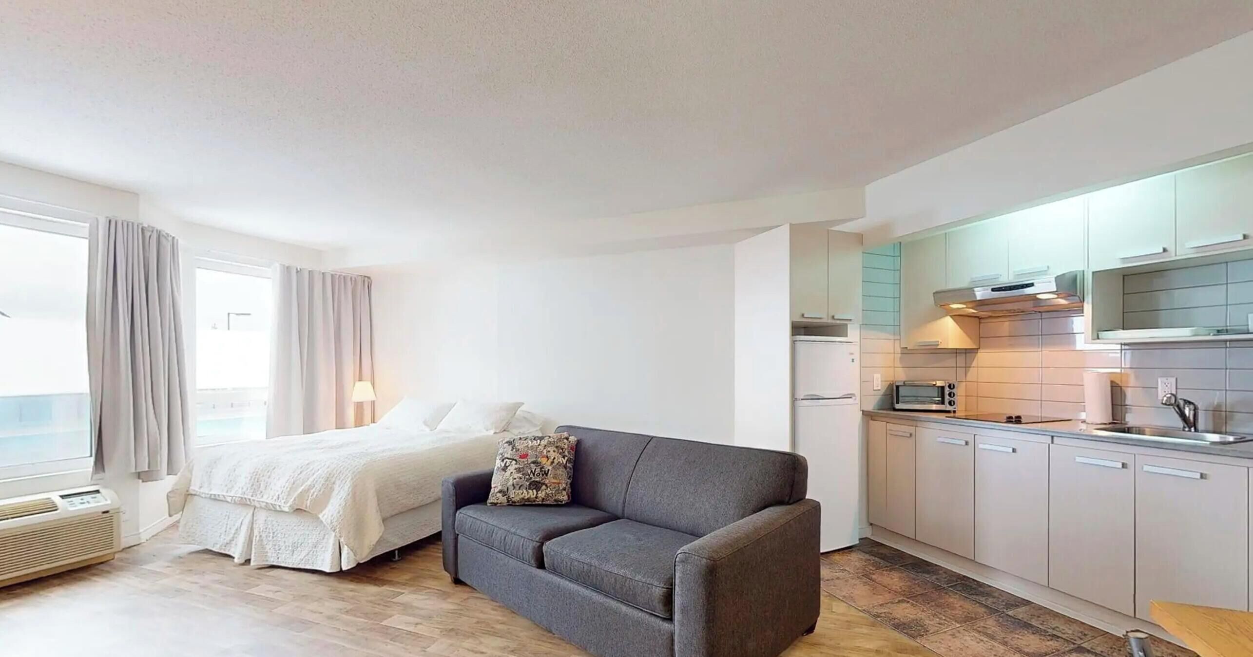 Ein kompaktes Hotelzimmer mit Bett, grauem Sofa, einer Küchenzeile und einem Fenster mit Vorhängen.
