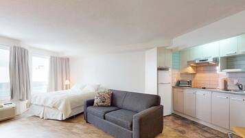 Ein kompaktes Hotelzimmer mit Bett, grauem Sofa, einer Küchenzeile und einem Fenster mit Vorhängen.