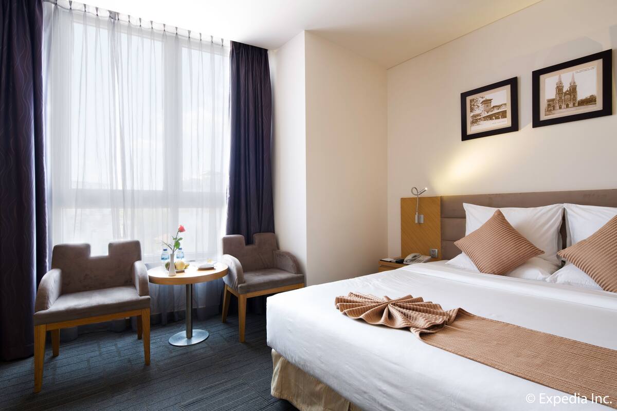 A hotel room with a bed, two chairs, a small table with a vase and a flower, and two framed pictures on the wall.