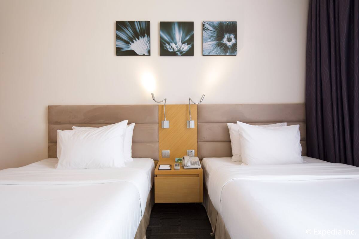 A hotel room with two beds, a wooden headboard, a nightstand with a phone and a lamp, and three wall-mounted artworks.