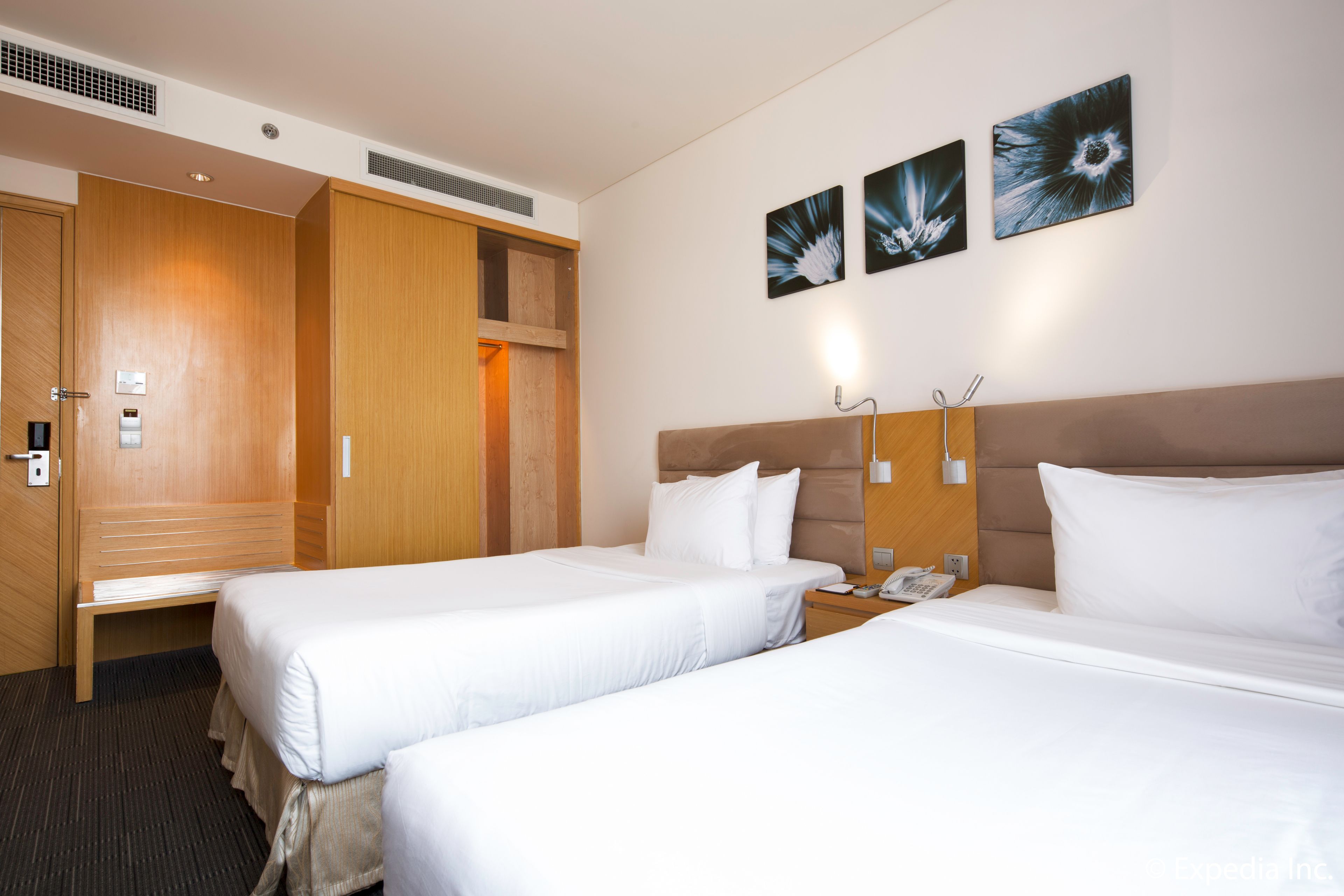 A hotel room with two beds, a wooden wardrobe, a bedside table with a phone, and wall-mounted artwork.