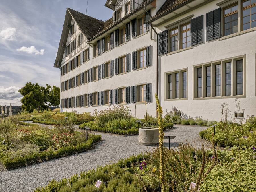 Un edificio de varios pisos con fachada simétrica, ventanas amplias y un sendero de grava que conduce a una jardinera de hormigón central.