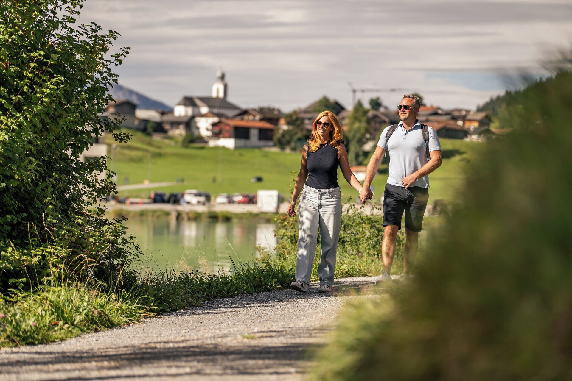 A couple walking on a gravel path with a body of water and houses in the background.