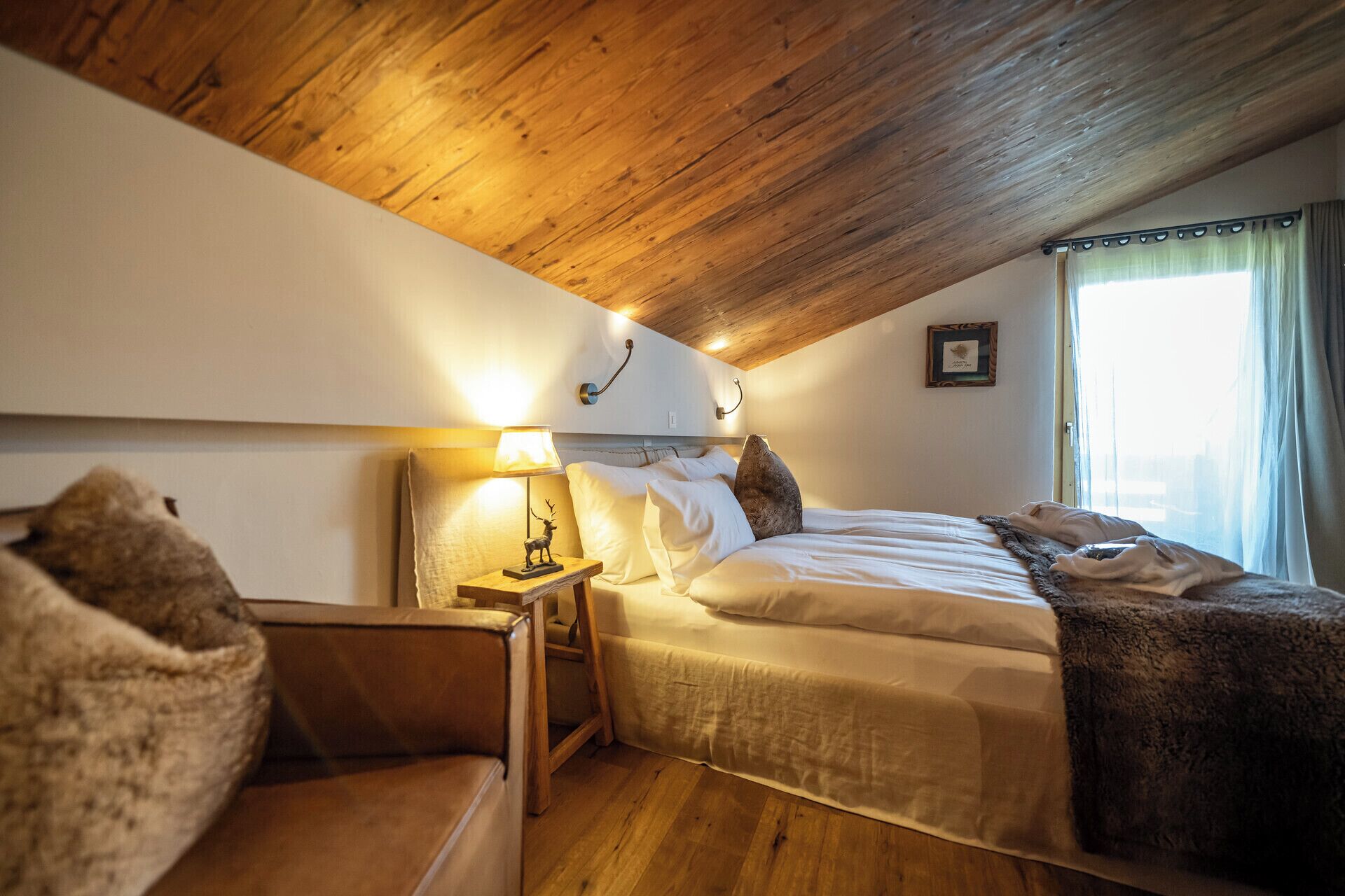 A cozy bedroom with a wooden ceiling, a bed with white linens, a nightstand with a lamp, and a window with curtains.