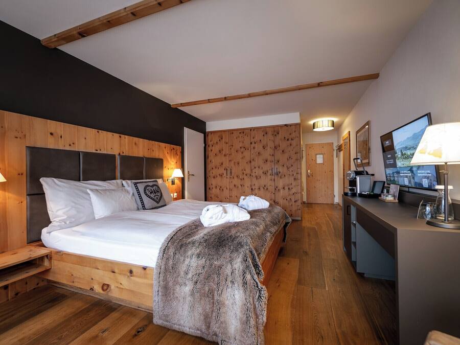 Una habitación de hotel moderna con cabecera de madera, una cama grande con ropa blanca, un cubrecama de piel, un escritorio con lámpara y un televisor que muestra una vista de montañas.