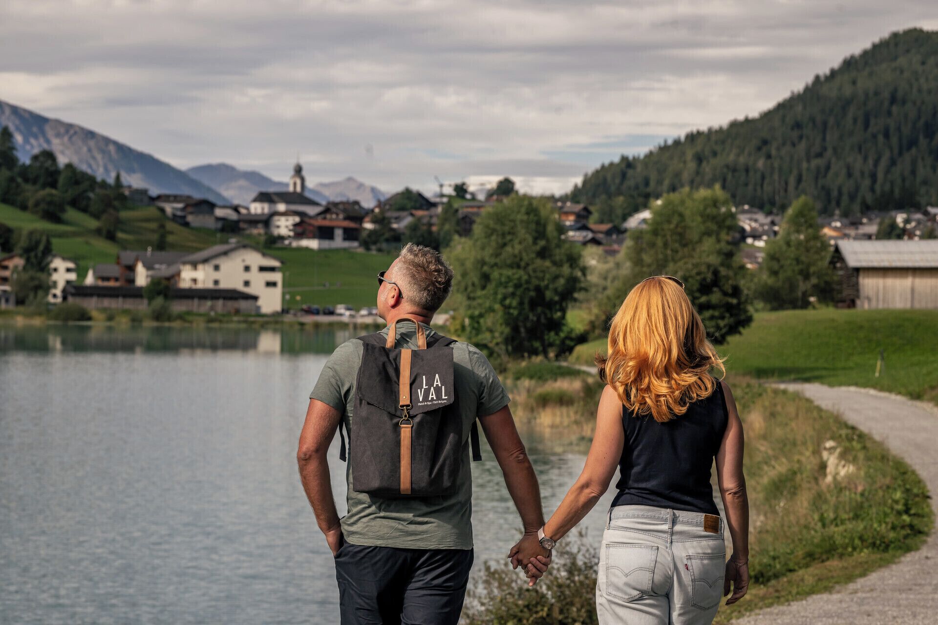 Dos personas tomadas de la mano frente a un lago, con casas y montañas al fondo.