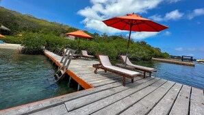 Une jetée en bois, agrémentée de chaises longues et de parasols rouges, s’avance sur des eaux calmes sous un ciel dégagé.