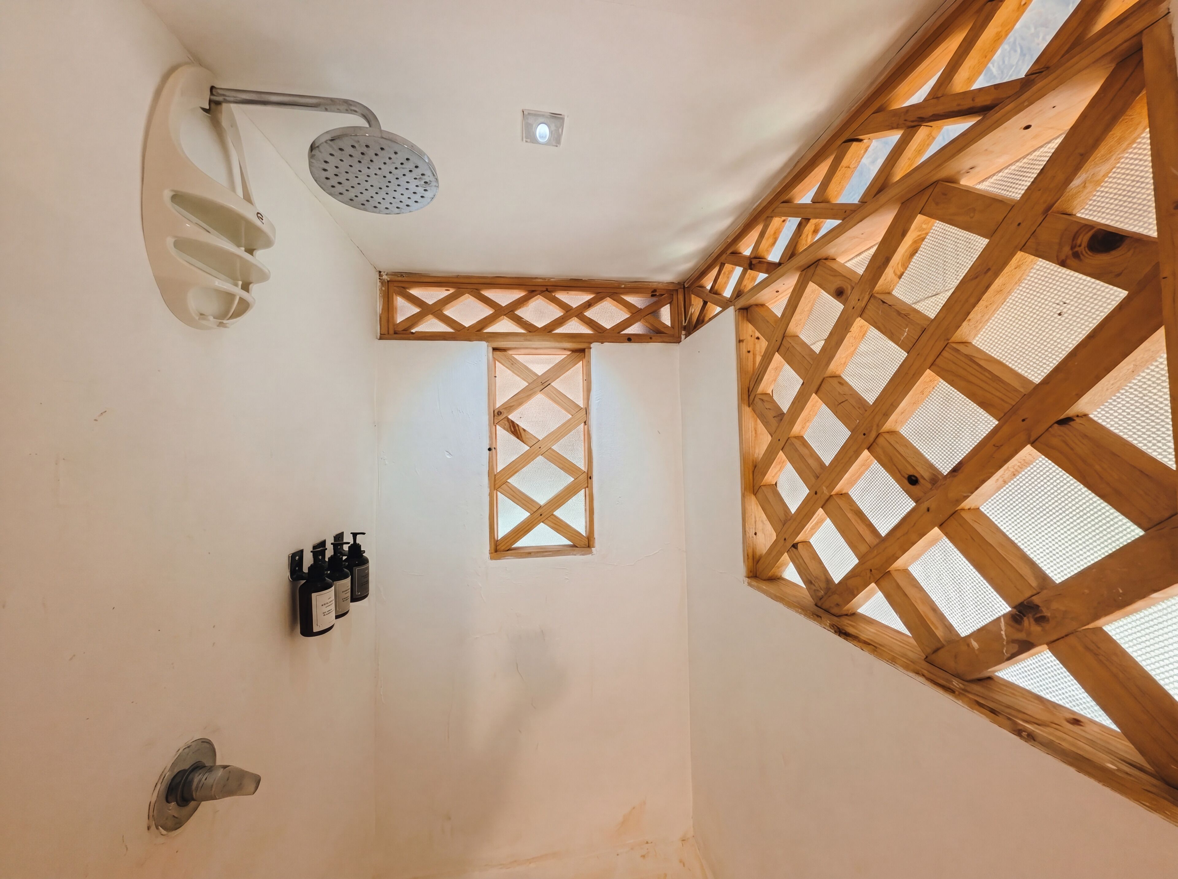 Una ducha con cabezal de ducha de mano, una ventana pequeña con diseño de celosía de madera y un dispensador de jabón instalado en la pared.
