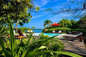 A pool area with lounge chairs and umbrellas, surrounded by lush greenery and overlooking a clear blue sea. - Hotel PlayaScondida Baru (Cartagena)