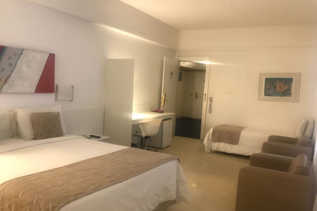 Une chambre d’hôtel avec un grand lit, un bureau, une chaise et un petit canapé. Une porte donne accès à une salle de bain et à un couloir.