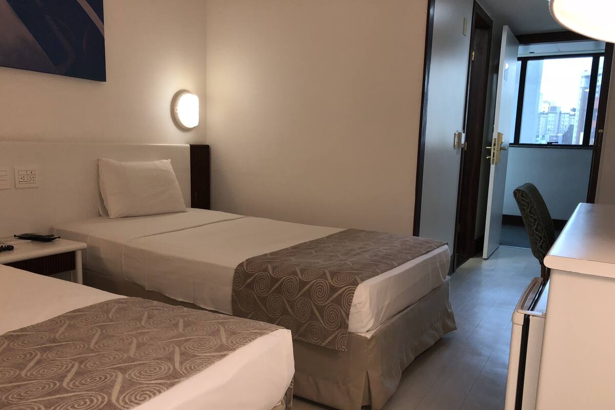 Une chambre d’hôtel avec deux lits, une table de chevet avec un téléphone et une télécommande, une lampe fixée au mur et une vue sur la ville par la fenêtre.