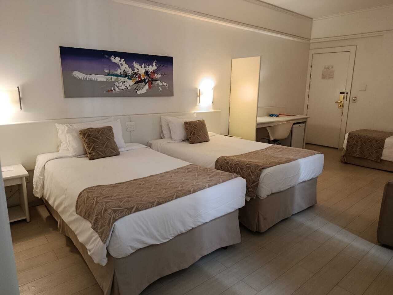 A hotel room with two beds, a desk, a chair, and a painting on the wall.