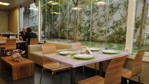 Um restaurante com decoração moderna, exibindo uma mesa para dois com jogos americanos verdes, cadeiras de madeira e um vaso de flores sobre uma mesa de madeira.