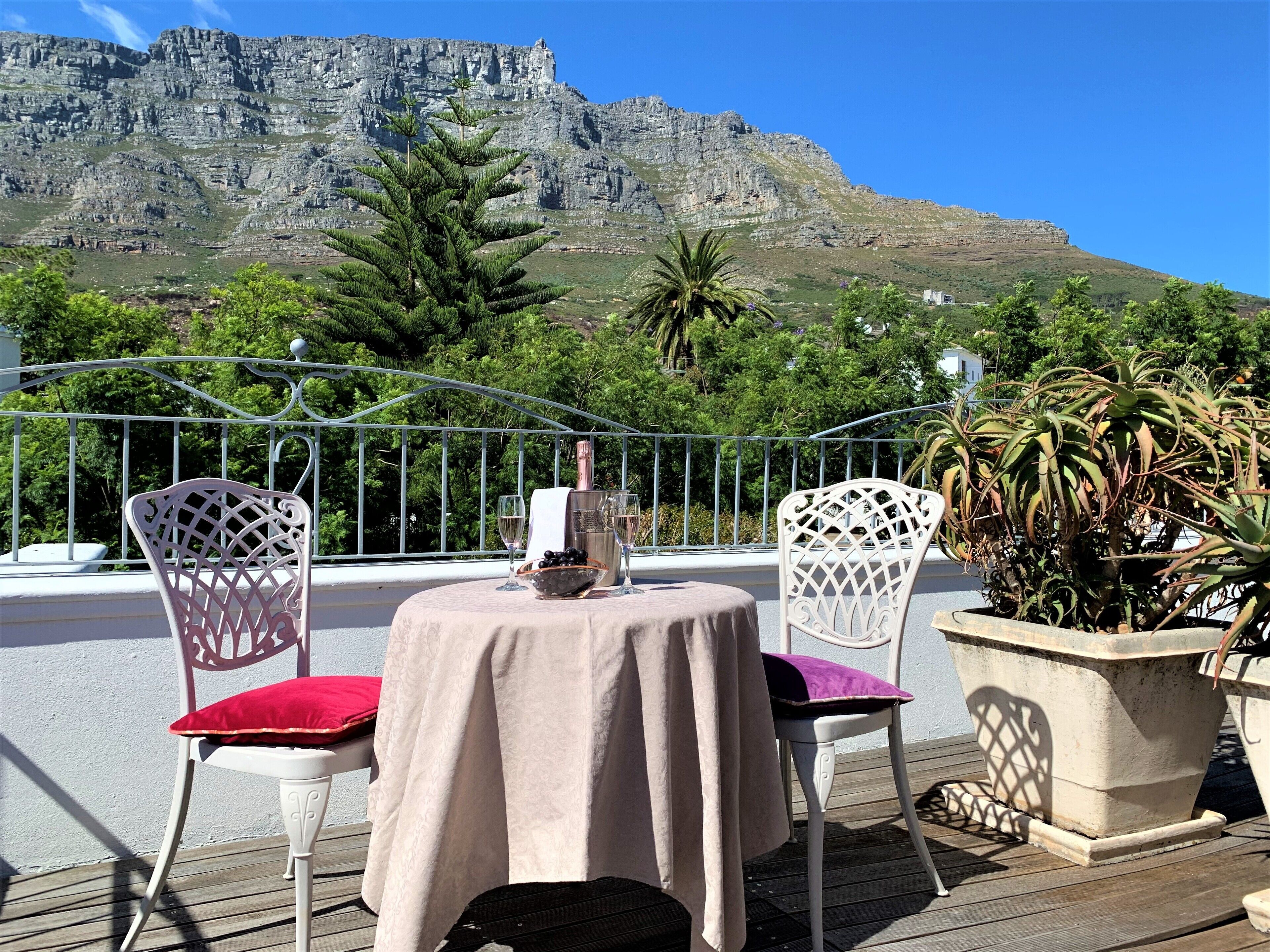 A table set with a bottle, glasses, and grapes on a balcony with chairs, overlooking a mountainous landscape.