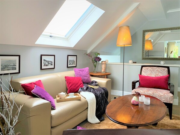 A cozy living room with a high ceiling, a skylight, a round wooden coffee table, a sofa with cushions, a chair, and a mirror on the wall.