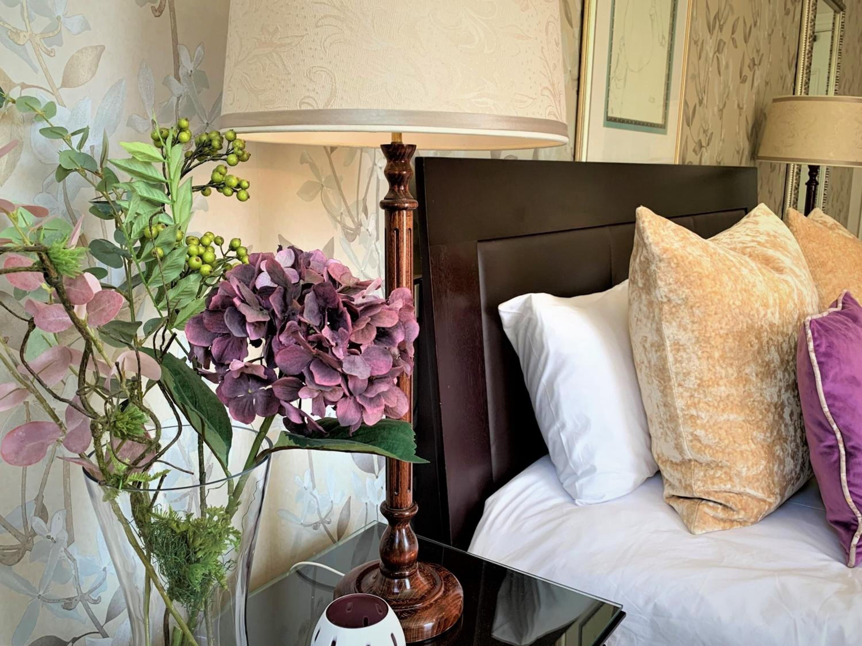 A neatly made bed with white and beige pillows, a dark headboard, and a bedside table with a lamp and a vase of purple flowers.