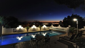 A poolside area with a view of the city at dusk, outdoor lighting, and lounge chairs.