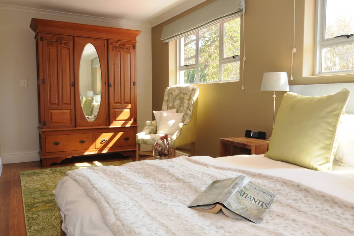 Une chambre avec une grande armoire en bois, un fauteuil à motifs floraux, une table de chevet avec une lampe et un lit sur lequel se trouve un magazine.