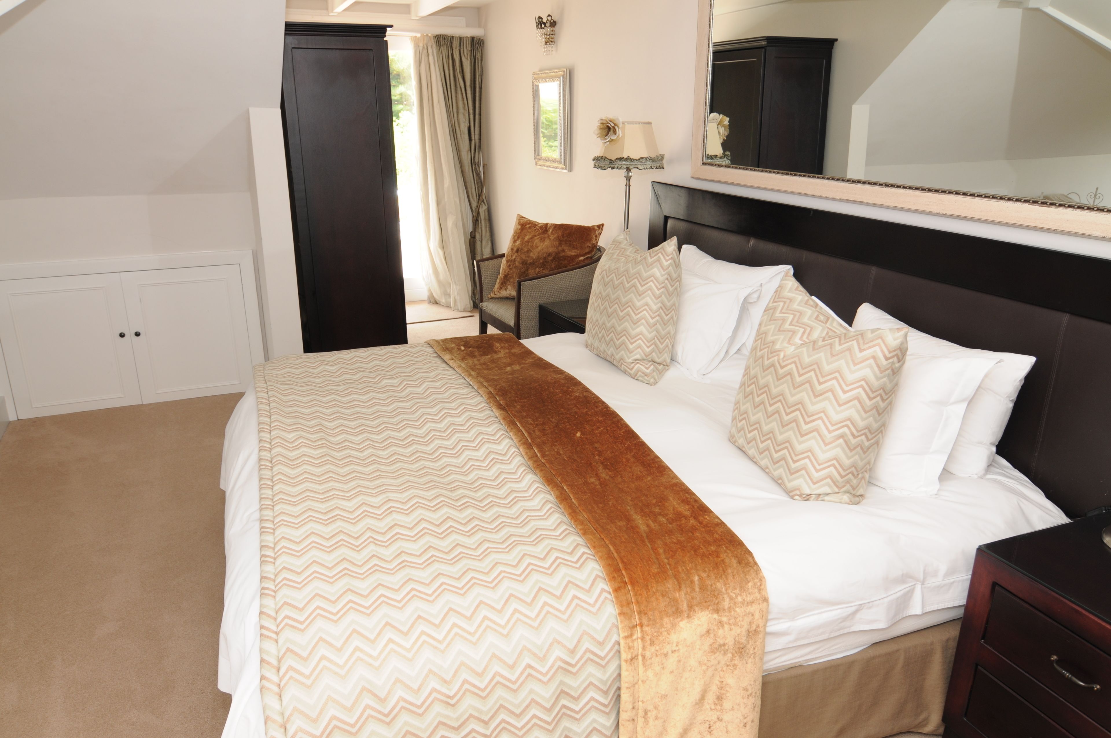 A neatly made bed with a patterned bedspread and a brown throw, a bedside table with a lamp, a chair with a cushion, and a wardrobe.