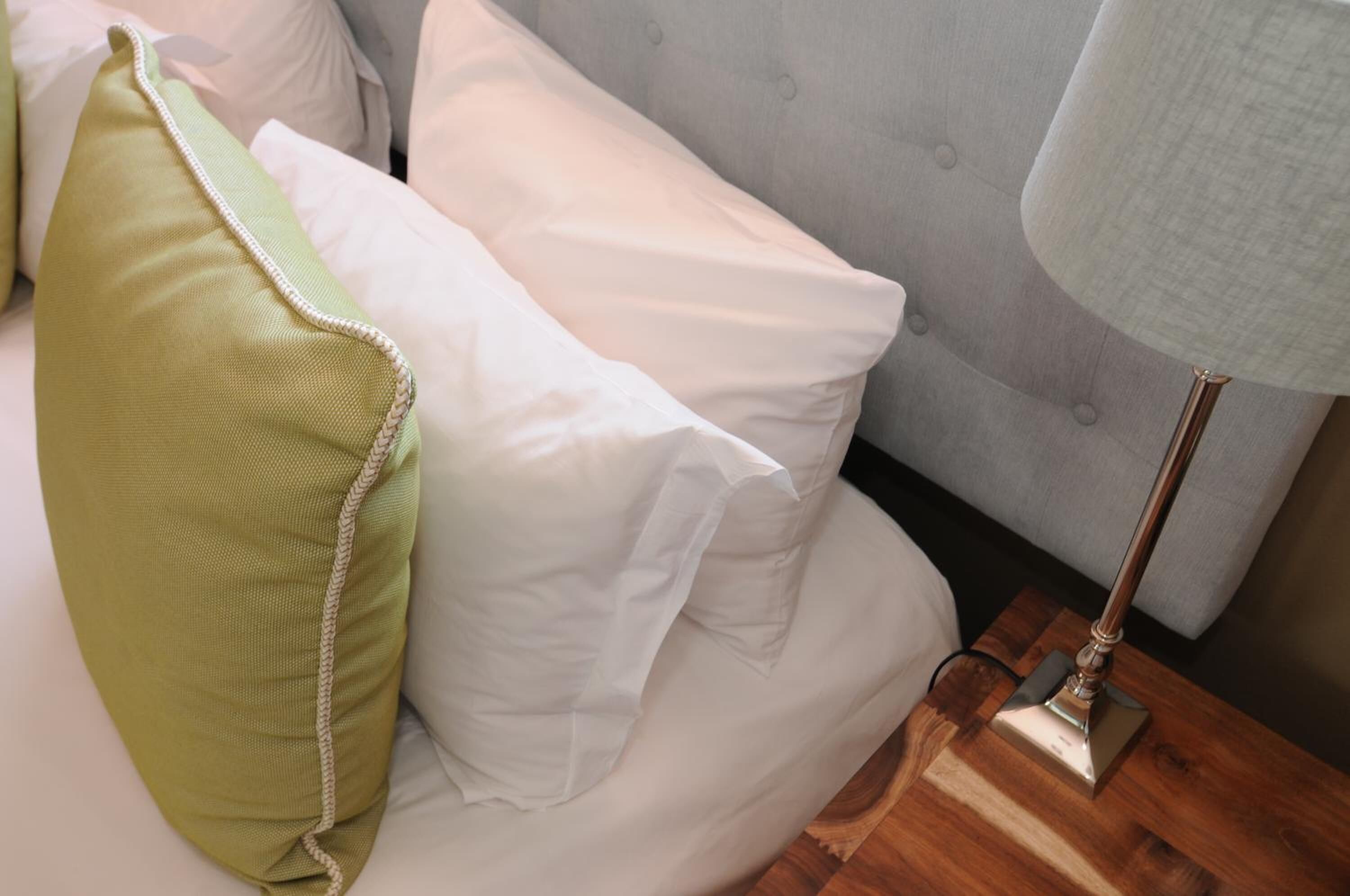 A bed with white and green pillows, a grey tufted headboard, and a wooden nightstand with a lamp.