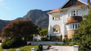 A two-story house with a stone base and white upper structure, featuring a balcony and a view of a mountain.
