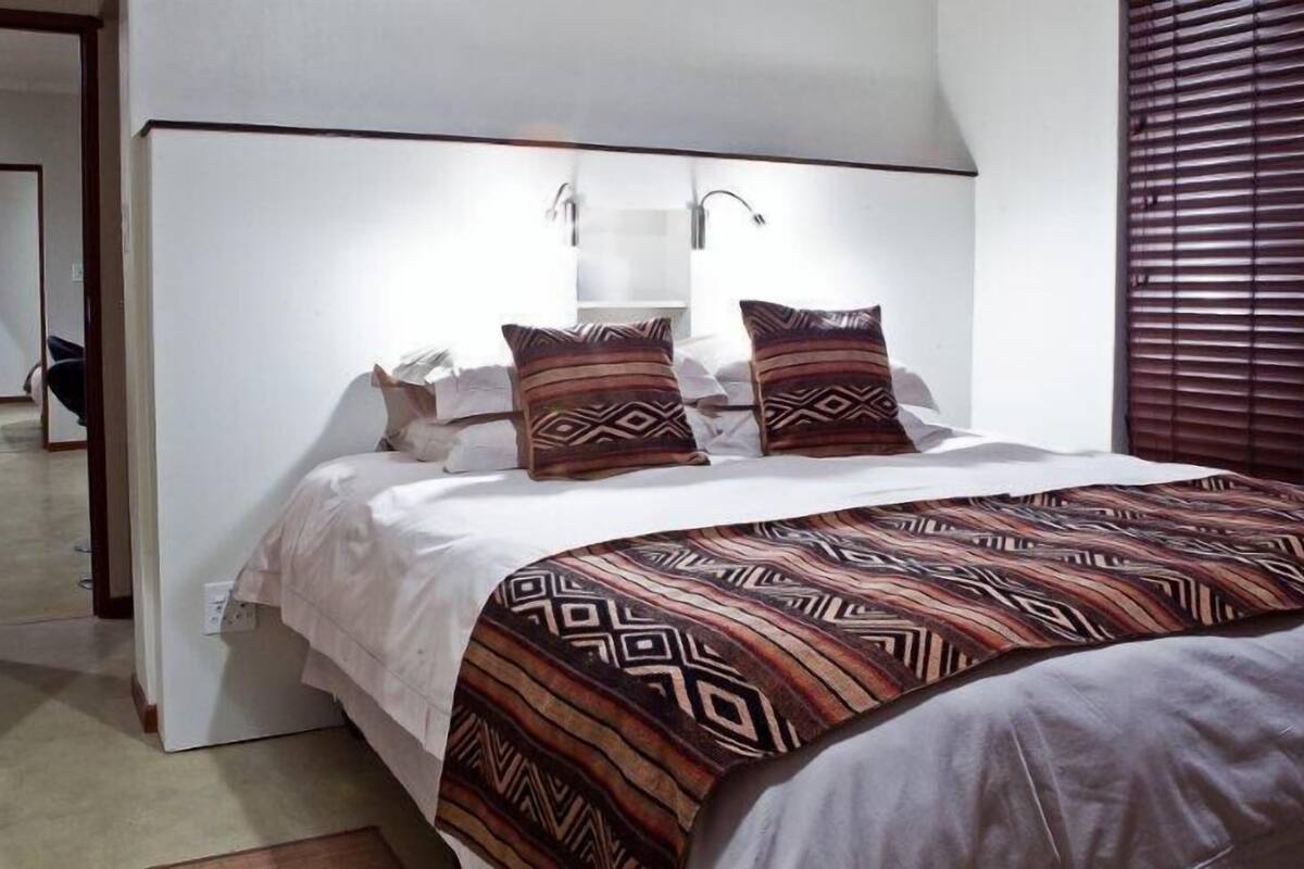 Een slaapkamer met een bed, kussens, een nachtkastje en een raam met jaloezieën.