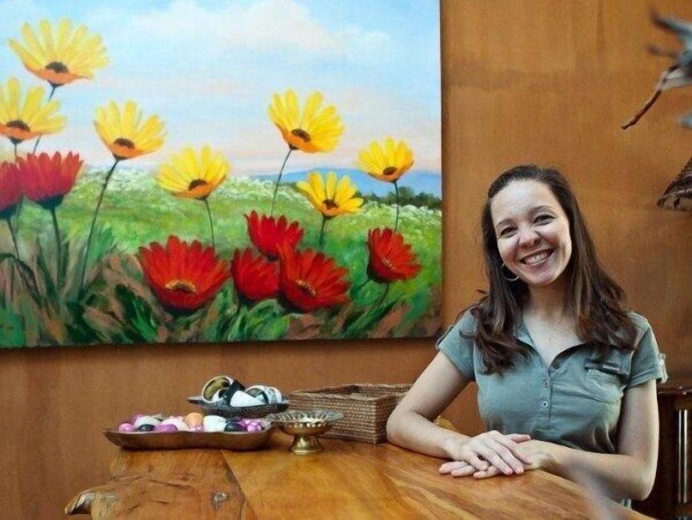 Une femme est assise à une table en bois, avec une peinture de fleurs en arrière-plan.